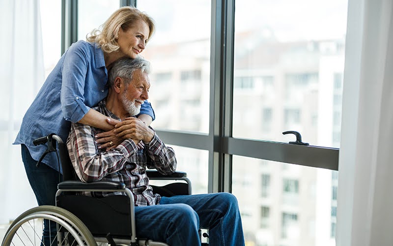 Senior woman and her husband in a wheelchair looking through window from their home. 